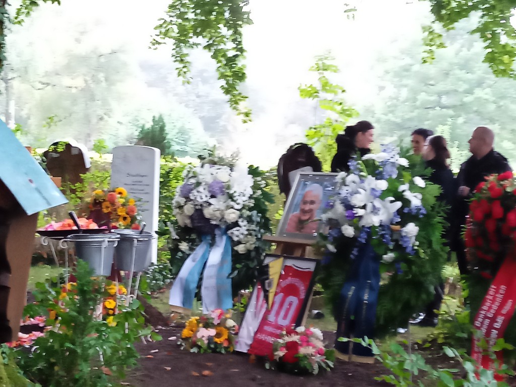 Grabfoto Klaus Hahnzog mit Portrait, Trikots, Blumenschmuck und Kränzen, Personen im Hintergrund